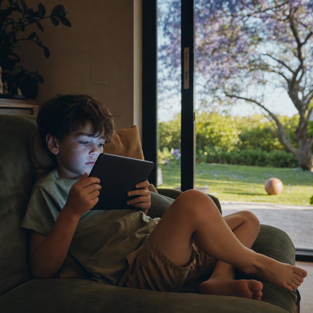 a boy on the couch lit up by a tablet while the sunny garden waits outside